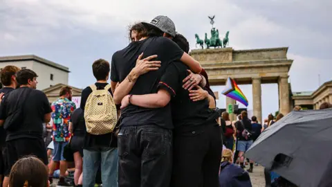 ผู้คนสามคนกอดกัน มีธง LGBT สีรุ้งและประตูบรันเดินบวร์กอยู่เบื้องหลัง