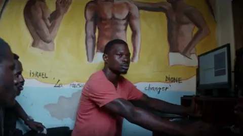 Artists Equity Rapper Stone uses a computer. Two men watch him.