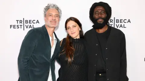 Getty Images for Tribeca Festival Taika Waititi, Dione Roach and Steve Happi pose for a photo in front of a Tribeca Festival step and repeat