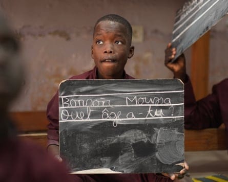 A young boy with a hopeful expression holds up a small blackboard with words in French written in chalk