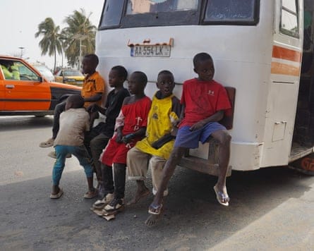 Five boys in dirty T-shirts mostly far too big for them sit on the rear bumper of a bus as another stands by them