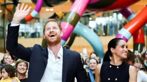 Reuters ด้านซ้าย เจ้าชายแฮร์รียิ้มแย้ม เงยหน้ามองและโบกมือขวาในอากาศ ด้านขวา เมแกนยิ้มและมองไปทางซ้ายของเธอ ท่ามกลางฝูงชนจำนวนมากและประติมากรรมสีสันสดใสขนาดใหญ่