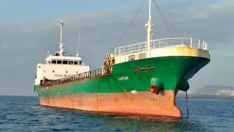 เรือ MV Lutuf จอดทอดสมออยู่ใกล้ชายฝั่ง มองเห็นตัวเรือสีเขียวและส้มเหนือผิวน้ำ