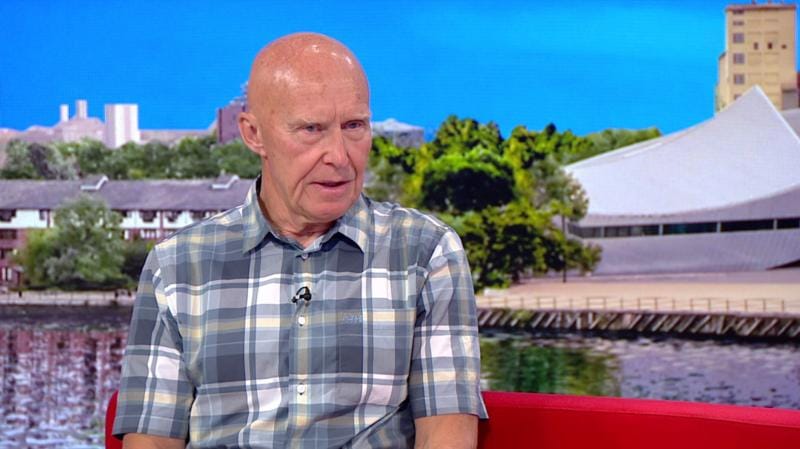 อลัน ฮินเคส สวมเสื้อลายตารางสีเทาขาว นั่งอยู่บนโซฟาสีแดงในสตูดิโอ BBC Breakfast