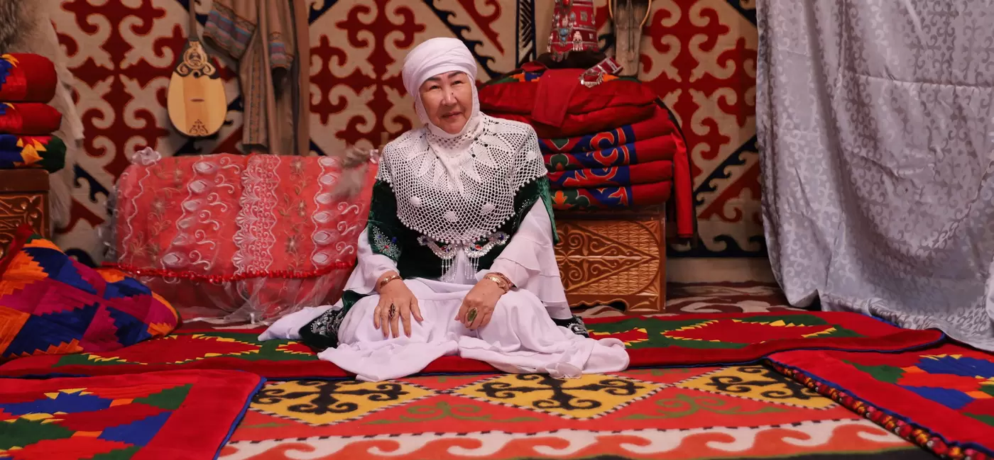 A woman in traditional Kazakh dress sits in a room draped with red patterned carpets