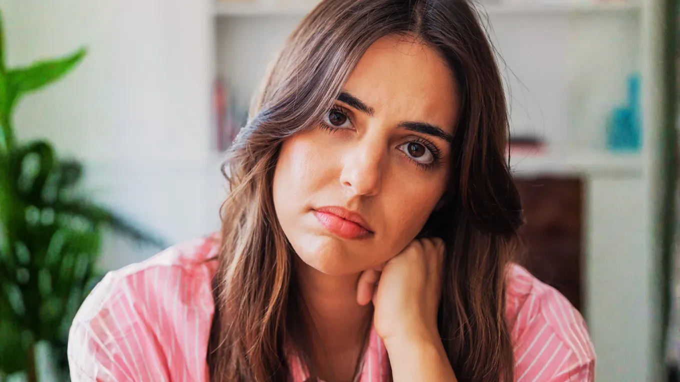 A young woman wearing a pink striped shirt leaning on her hand with a contemplative, guarded expression, symbolizing social manipulation, deceptive cues, and psychological self-defense.