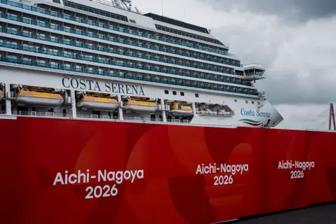 AFP via Getty Images เรือสำราญคอสตา เซเรนา (Costa Serena) จอดอยู่ท่ามกลางท้องฟ้าครึ้ม พร้อมป้ายขนาดใหญ่เขียนว่า Aichi-Nagoya 2026