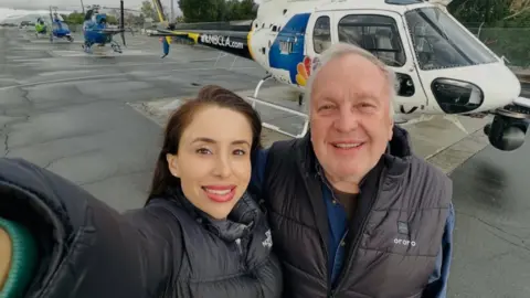 George Marciniw via CBS News ช่างภาพข่าวเอเลียนา โมเรโนและนักบินจอร์จ มาร์ชินิวถ่ายเซลฟี่ เธอสวมแจ็กเก็ตสีดำและยิ้ม เขาสวมเสื้อกั๊กสีดำและยิ้ม ทั้งคู่ยืนอยู่หน้าเฮลิคอปเตอร์บนพื้น