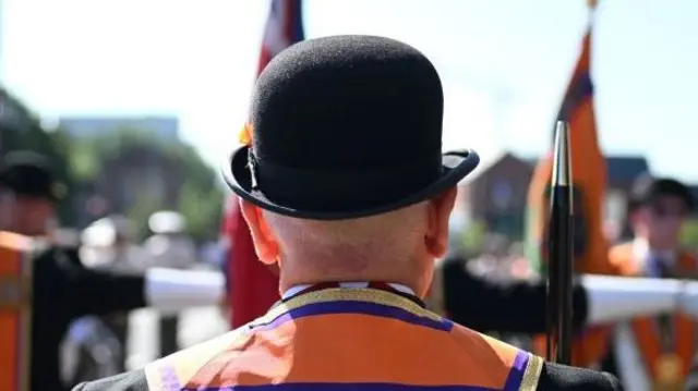 A man wears a black bowler hat, suit jacket and orange sash. We can only see the back of his head.